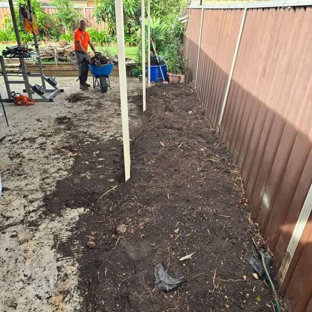 A worker in an orange shirt using a wheelbarrow on a cleared garden area.